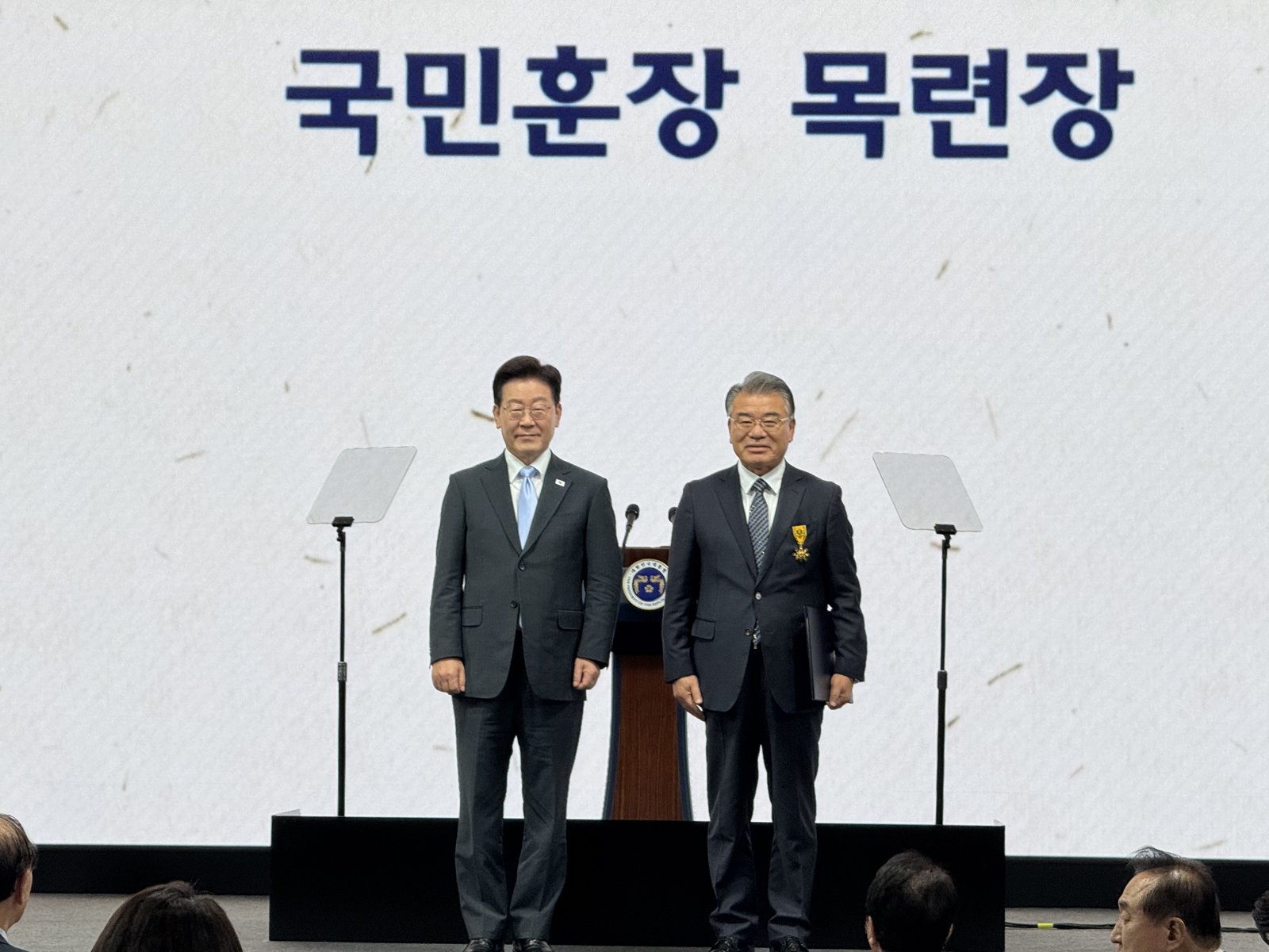 국민훈장 목련장 글자가 띄워진 무대에서 이종삼 갈릴리사랑의집 대표이사가 훈장을 달고 시상자와 나란히 서 있는 모습