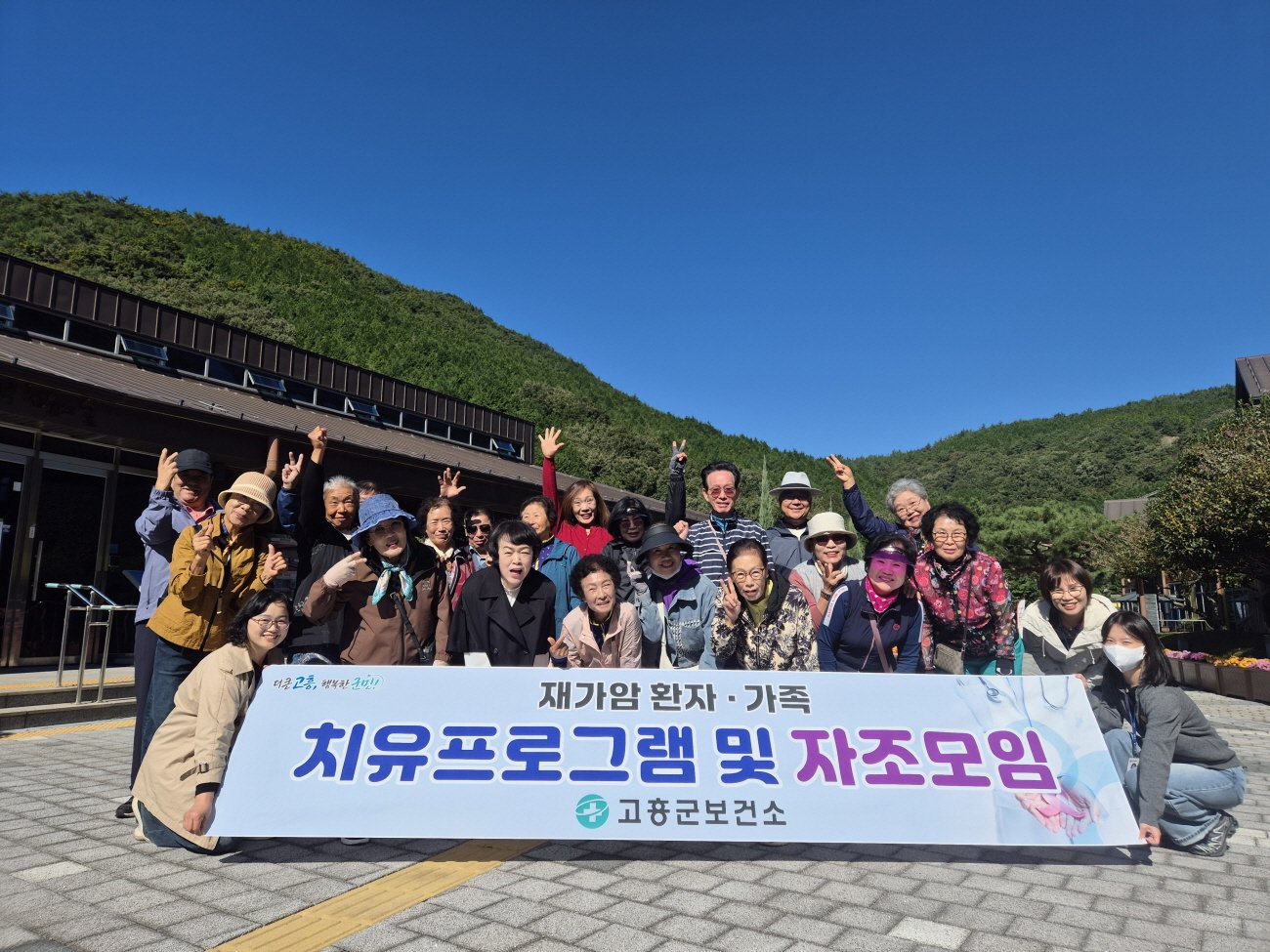 산자락 아래 건물 앞에서 참가자 20여 명이 '재가암 환자·가족 치유프로그램 및 자조모임'이라고 적힌 현수막을 들고 손을 들어 올린 채 웃고 있는 모습