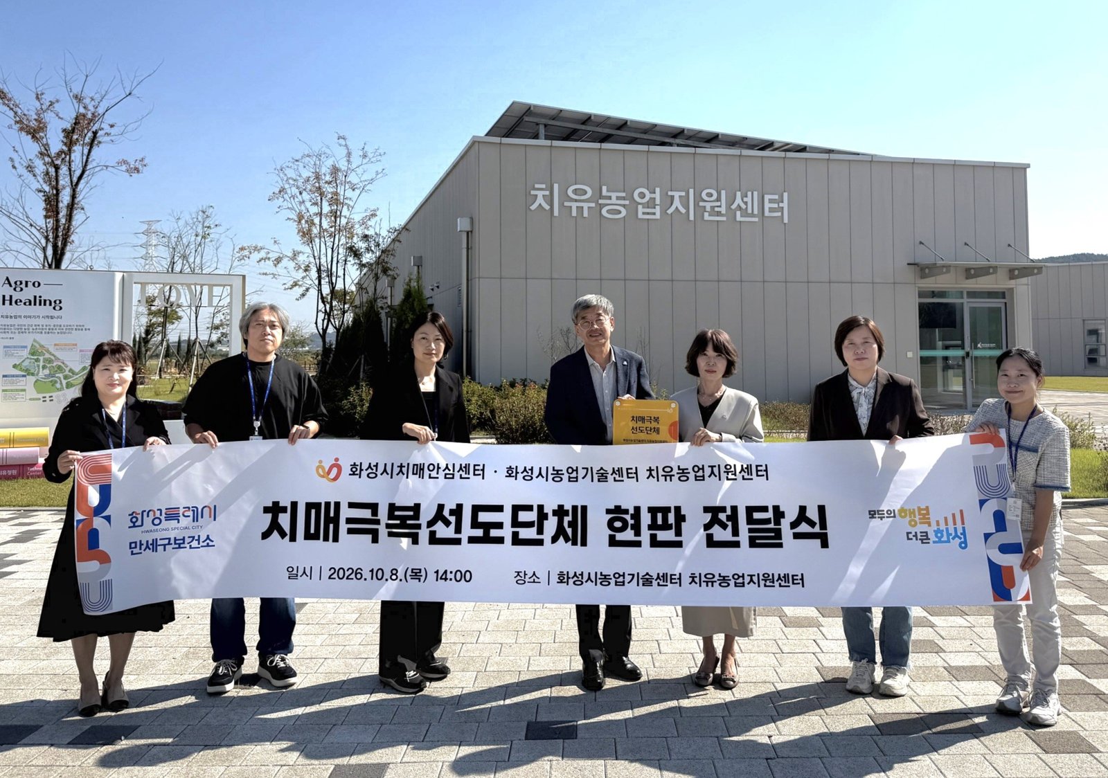화성시농업기술센터 치유농업지원센터 건물 앞에서 참석자 10여 명이 '치매극복선도단체 현판 전달식'이라고 적힌 현수막을 들고 서 있는 모습