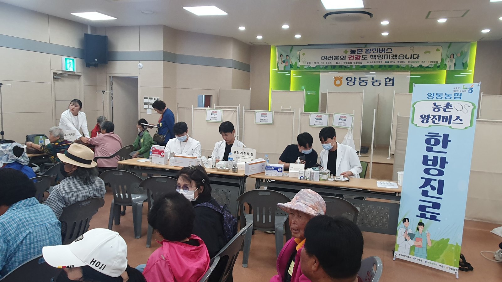 '한방진료' 안내 배너가 세워진 진료석 앞에서 흰 가운을 입은 의료진이 어르신들을 진료하는 모습