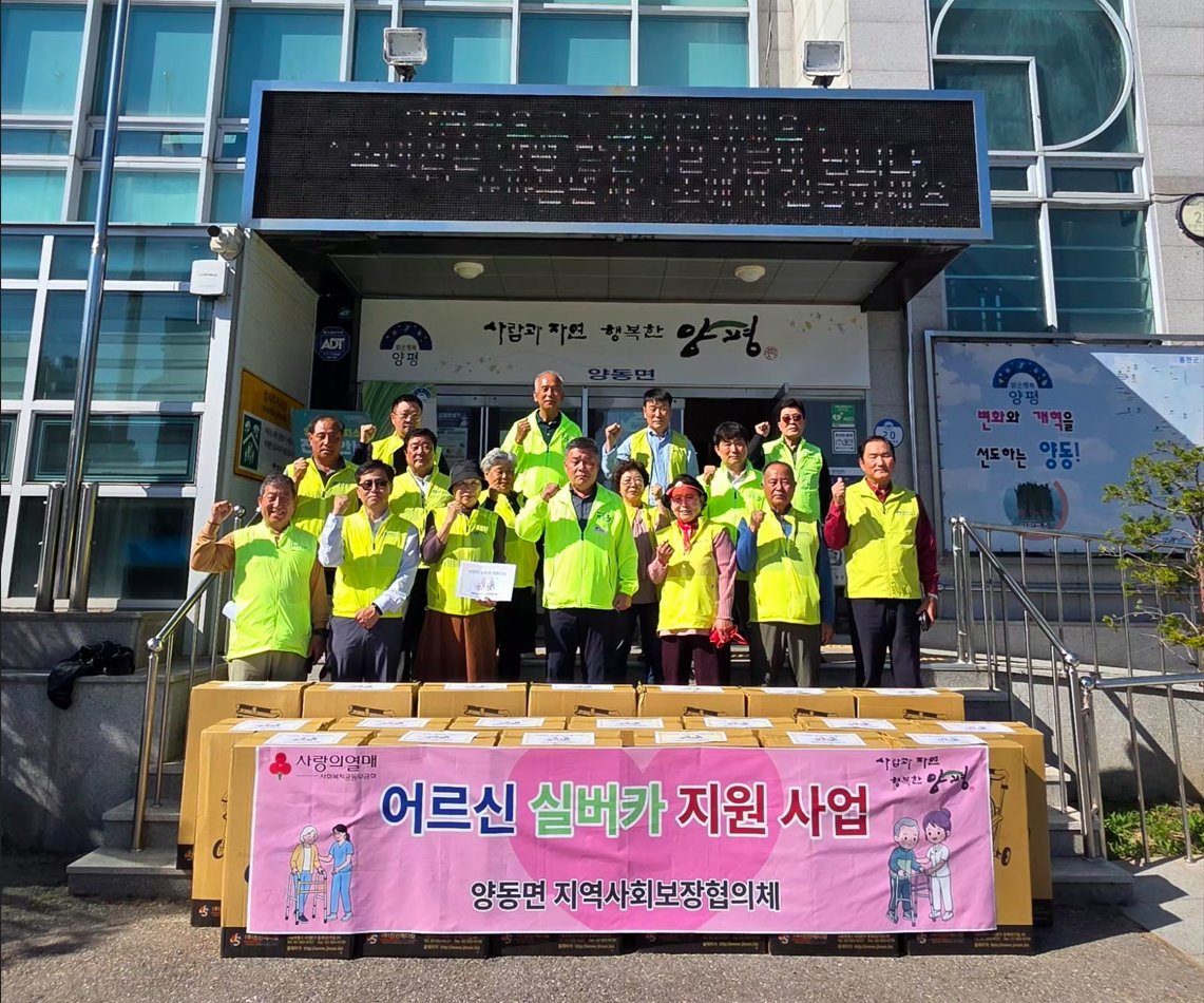 양동면 행정복지센터 앞에서 형광 조끼를 입은 협의체 위원들이 '어르신 실버카 지원 사업' 현수막과 실버카 상자들 뒤에 서서 손을 들고 있는 모습