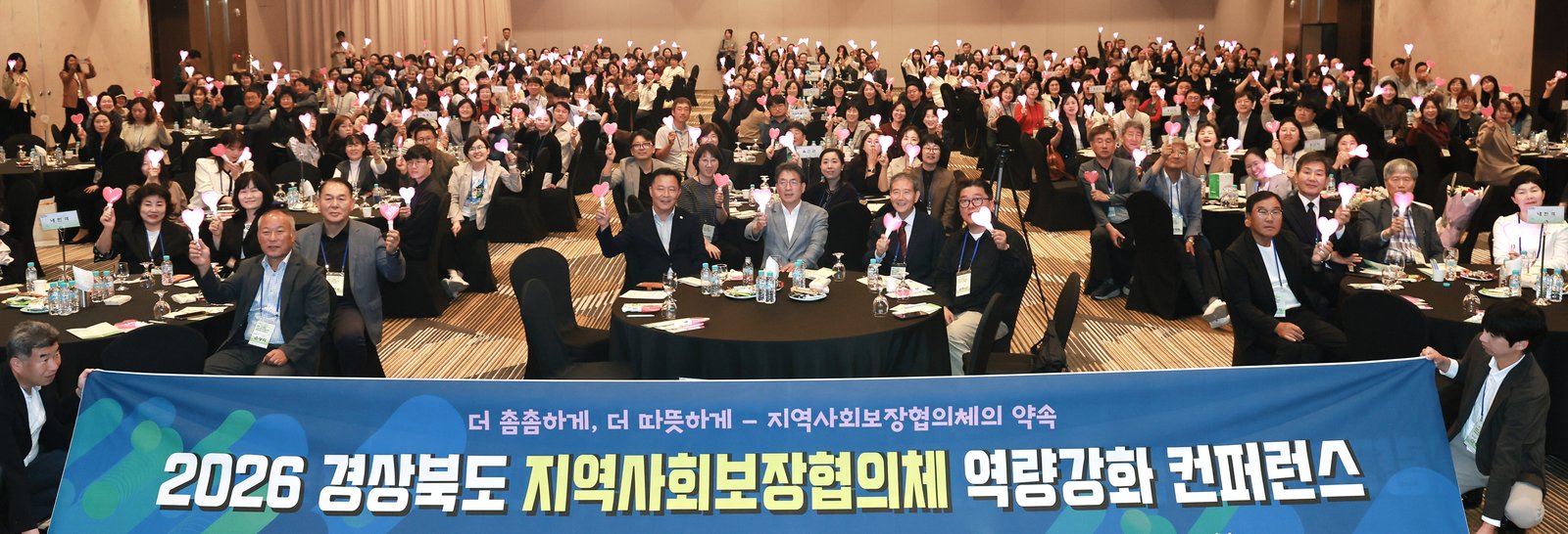 넓은 연회장 원탁에 앉은 경상북도 지역사회보장협의체 관계자들이 하트 모양 응원봉을 들고 있고, 앞줄에서 '2026 경상북도 지역사회보장협의체 역량강화 컨퍼런스' 현수막을 펼쳐 든 모습