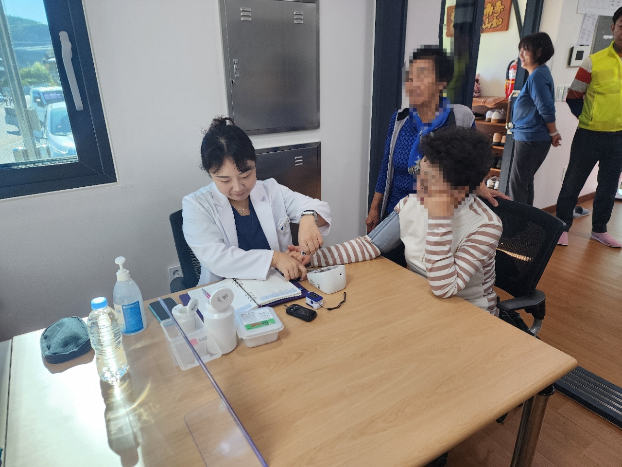 경로당 탁자에서 흰 가운을 입은 보건 인력이 의자에 앉은 어르신의 팔에 혈압계를 감고 측정하는 모습