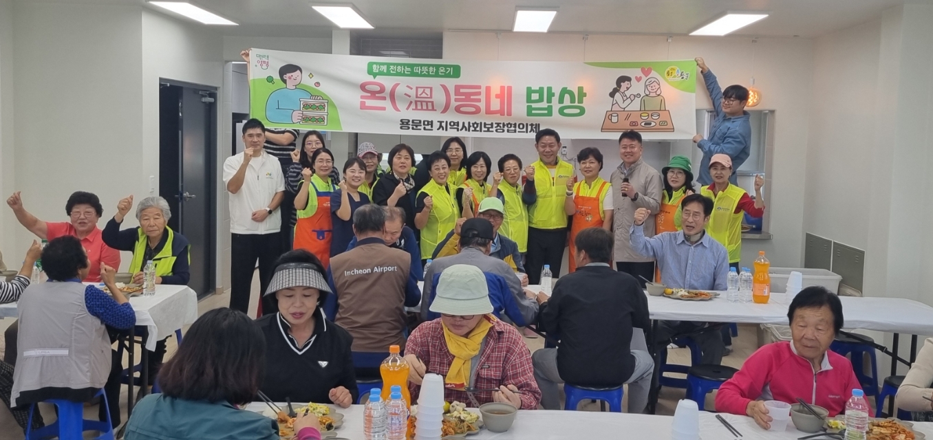 덕촌2리 경로당에서 '온(溫) 동네 밥상 용문면 지역사회보장협의체' 현수막 아래 형광 조끼를 입은 위원들이 주먹을 들어 보이고, 앞쪽 식탁에서 어르신들이 식사하는 모습