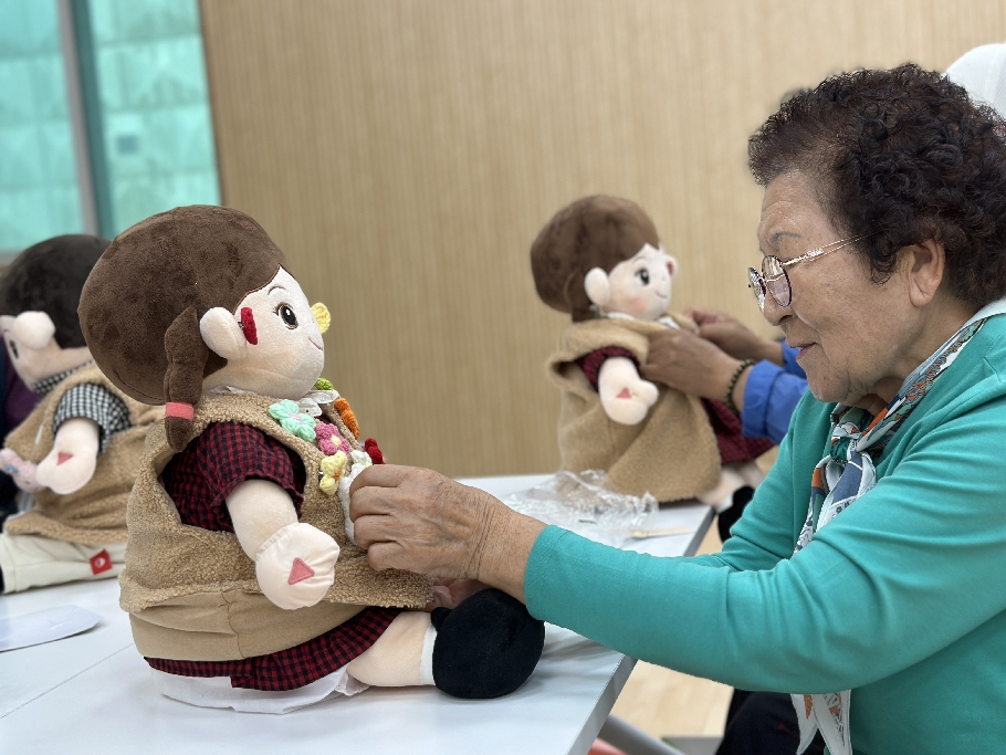 포천시 자조모임에서 연두색 옷을 입은 어르신이 탁자 위 인형 모양 인공지능 돌봄로봇에 꽃 장식을 달아 꾸미고 있다